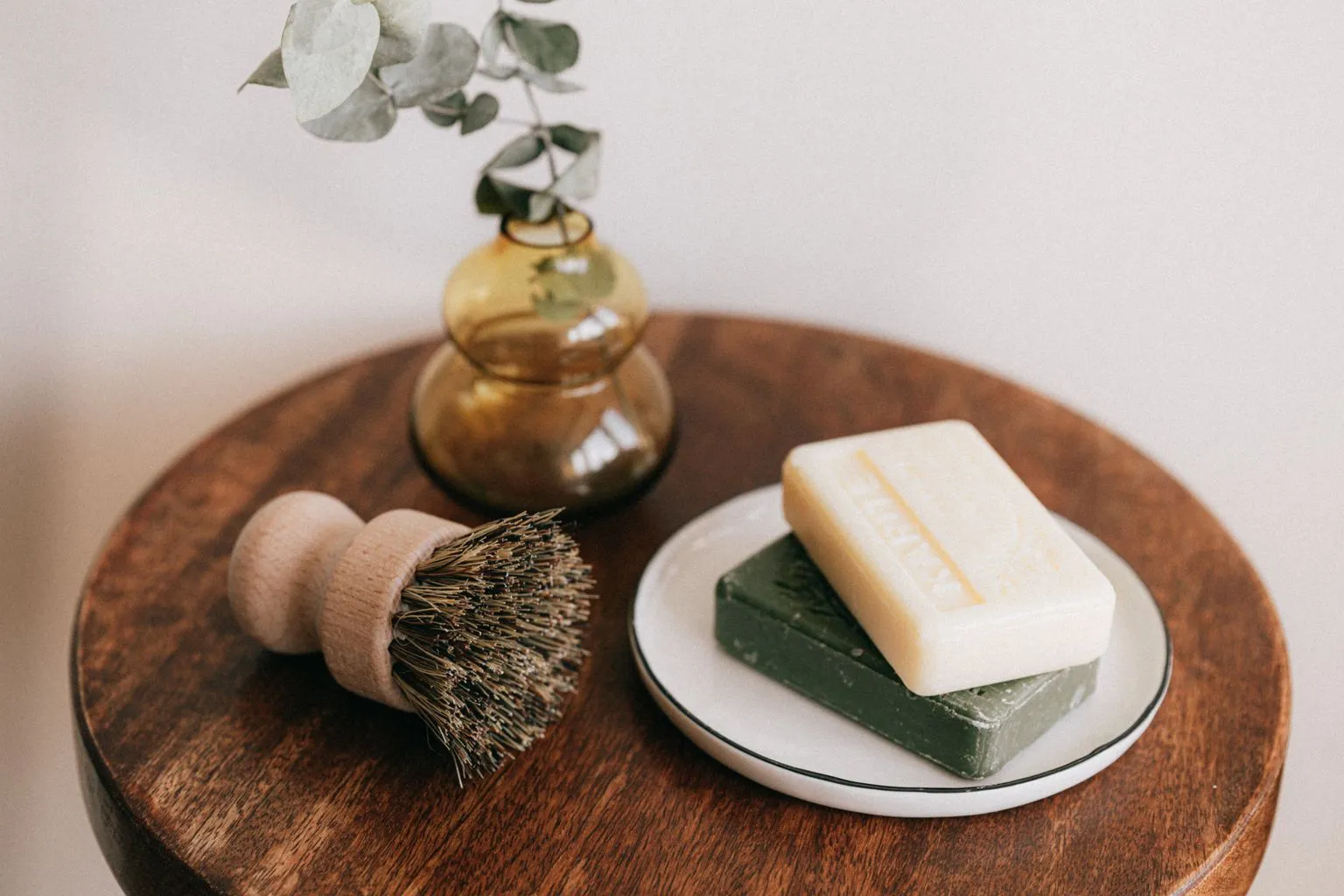 Two bars of soap on a ceramic dish beside a wooden dish brush and an amber glass vase of eucalyptus on a round wooden table.